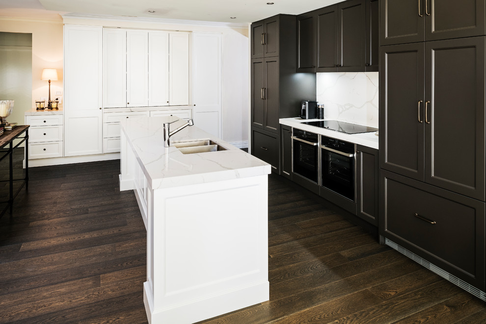 Photo of a large transitional galley open plan kitchen in Melbourne with a double-bowl sink, recessed-panel cabinets, brown cabinets, quartz benchtops, white splashback, stone slab splashback, black appliances, dark hardwood floors, with island, brown floor and white benchtop.