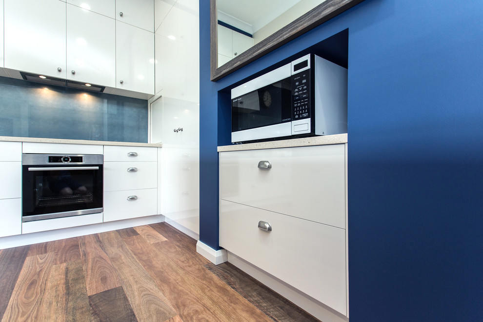 Inspiration for a small contemporary galley open plan kitchen in Other with a drop-in sink, flat-panel cabinets, white cabinets, solid surface benchtops, blue splashback, glass sheet splashback, stainless steel appliances, medium hardwood floors, a peninsula, brown floor and white benchtop.