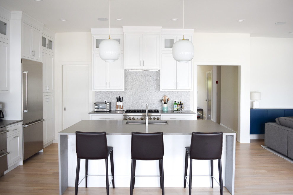 This is an example of a mid-sized beach style l-shaped eat-in kitchen in New York with an undermount sink, recessed-panel cabinets, white cabinets, quartz benchtops, grey splashback, marble splashback, stainless steel appliances, light hardwood floors and with island.