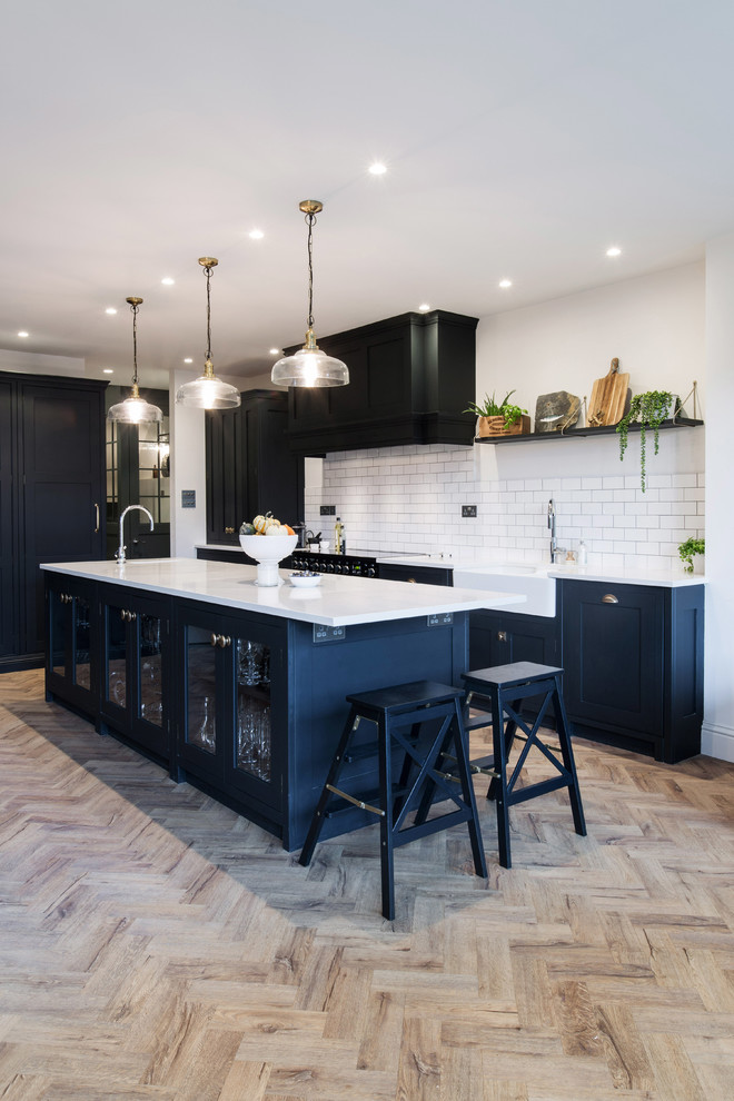 Photo of a large industrial galley eat-in kitchen in London with a farmhouse sink, glass-front cabinets, blue cabinets, granite benchtops, white splashback, ceramic splashback, black appliances, with island and white benchtop.