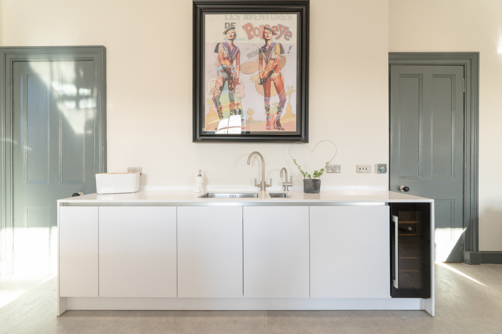 Photo of a large shabby-chic style l-shaped open plan kitchen in Edinburgh with an integrated sink, flat-panel cabinets, yellow cabinets, quartz worktops, beige splashback, stone tiled splashback, stainless steel appliances, cement flooring, an island, beige floors and white worktops.