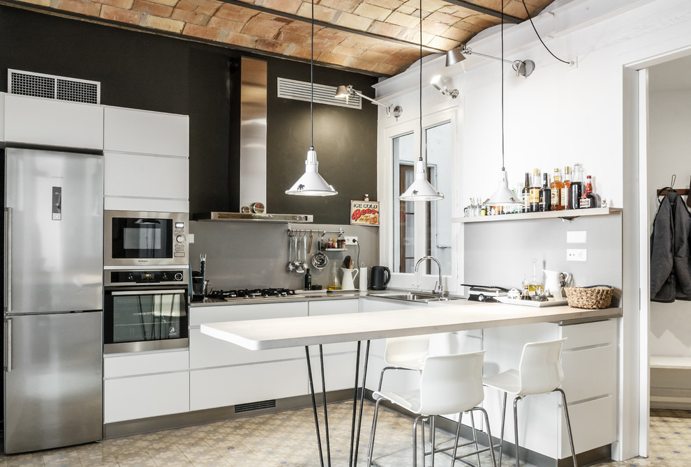 Photo of a mid-sized industrial l-shaped kitchen in Barcelona with flat-panel cabinets, white cabinets, grey splashback, ceramic floors, a peninsula and stainless steel appliances.