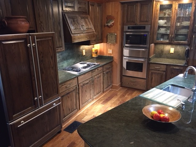 Large mountain style l-shaped medium tone wood floor and brown floor kitchen photo with an undermount sink, raised-panel cabinets, dark wood cabinets, granite countertops, stainless steel appliances and an island