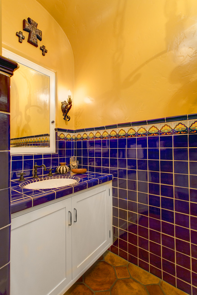 Photo of a mid-sized mediterranean powder room in Los Angeles with shaker cabinets, white cabinets, blue tile, multi-coloured tile, ceramic tile, yellow walls, terra-cotta floors, an undermount sink, tile benchtops, red floor and blue benchtops.