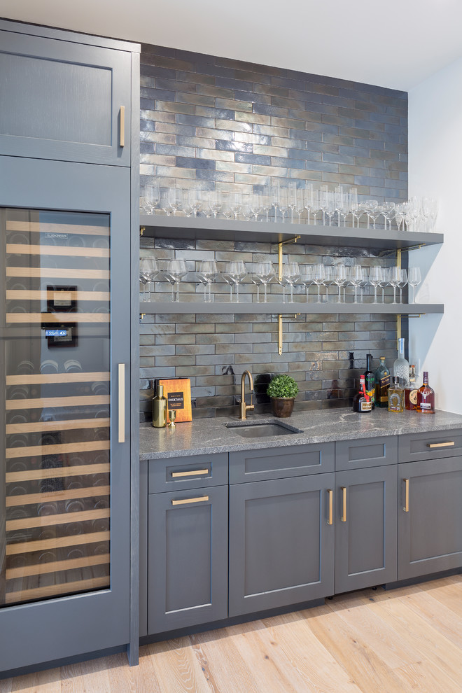 Design ideas for a large contemporary single-wall wet bar in Grand Rapids with an undermount sink, shaker cabinets, grey cabinets, granite benchtops, brick splashback, light hardwood floors, brown floor and grey benchtop.