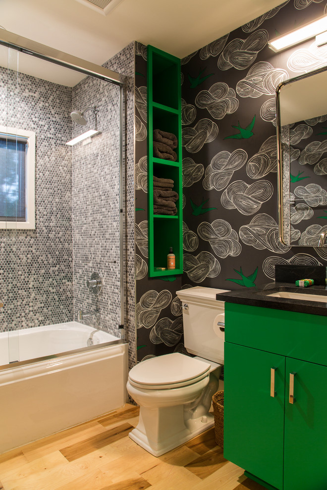 Contemporary bathroom in Austin with flat-panel cabinets, green cabinets, an alcove tub, a shower/bathtub combo, a two-piece toilet, black tile, gray tile, white tile, mosaic tile, black walls, light hardwood floors and an undermount sink.