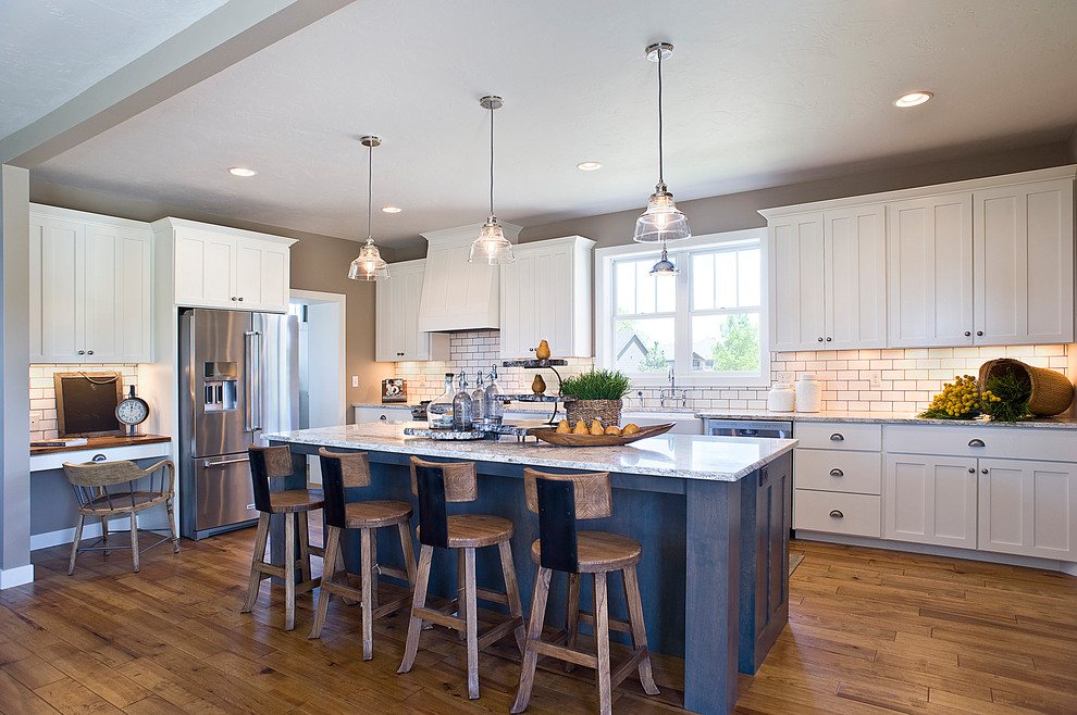 Example of a mid-sized transitional l-shaped medium tone wood floor and brown floor eat-in kitchen design with a farmhouse sink, raised-panel cabinets, white cabinets, granite countertops, white backsplash, subway tile backsplash, stainless steel appliances and an island