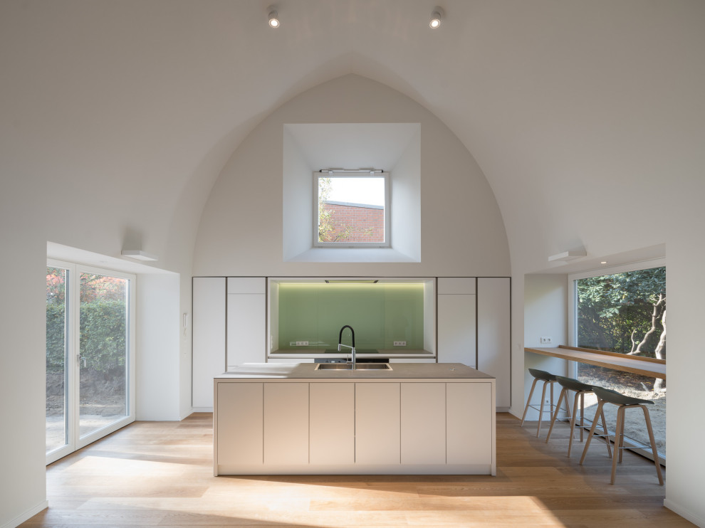 Mid-sized modern single-wall open plan kitchen in Hamburg with a double-bowl sink, flat-panel cabinets, white cabinets, laminate benchtops, green splashback, glass tile splashback, stainless steel appliances, medium hardwood floors, with island, brown floor, grey benchtop and vaulted.