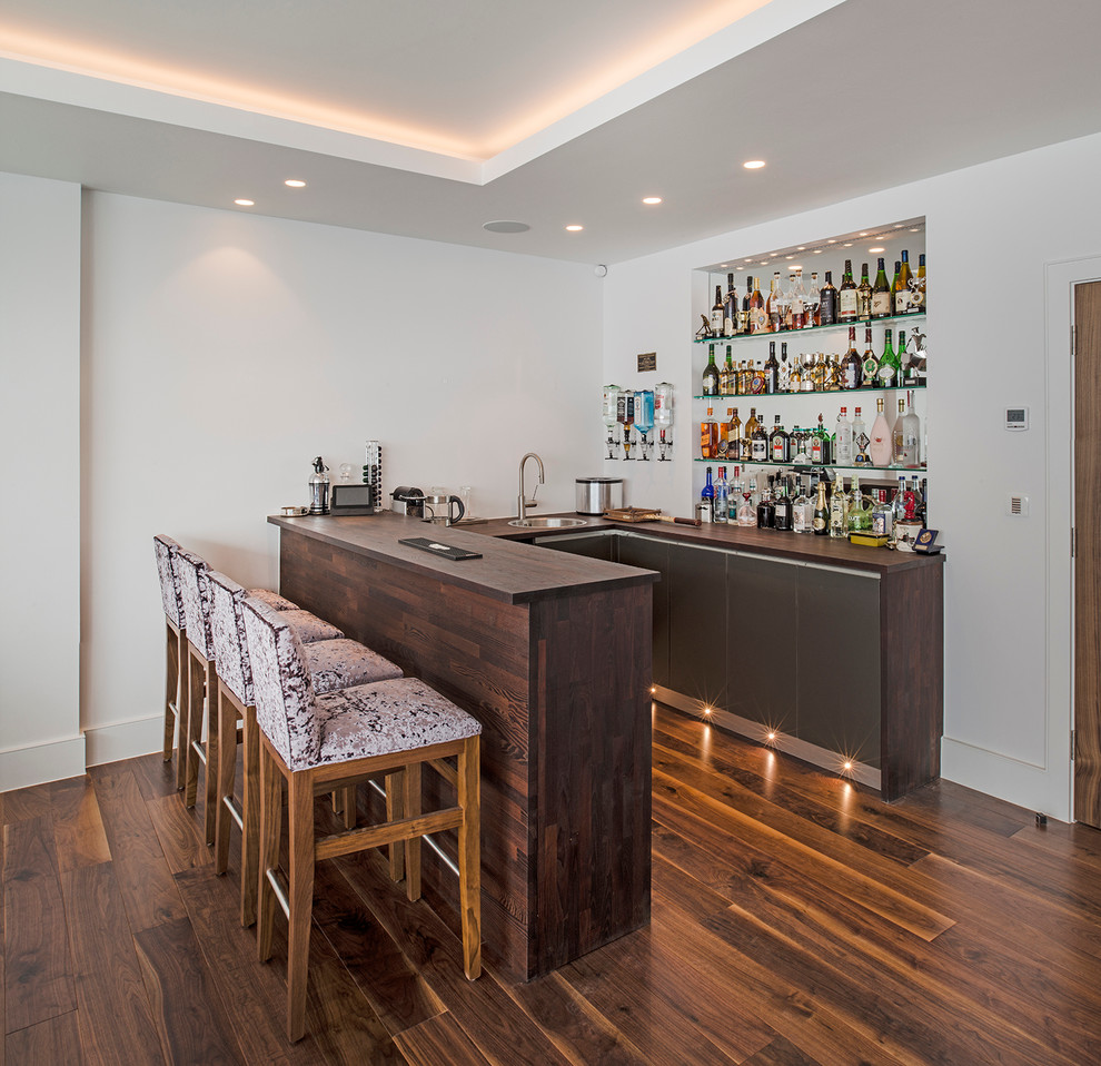 Photo of a contemporary u-shaped seated home bar in London with flat-panel cabinets, wood benchtops, medium hardwood floors and brown benchtop.