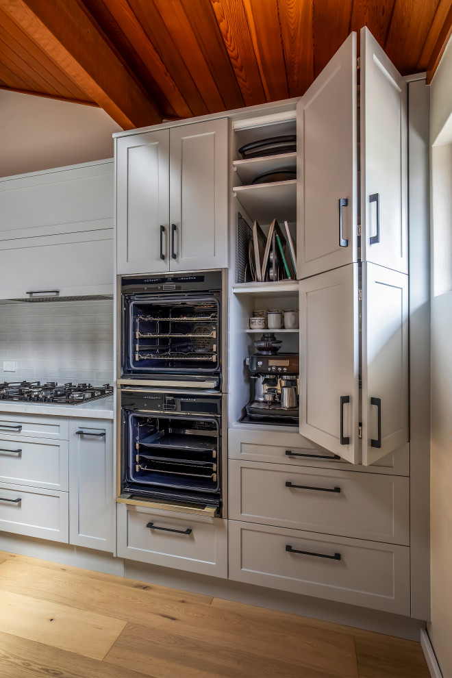 Large transitional galley eat-in kitchen in Sydney with a drop-in sink, shaker cabinets, white cabinets, quartz benchtops, white splashback, porcelain splashback, black appliances, light hardwood floors, with island, white benchtop and vaulted.