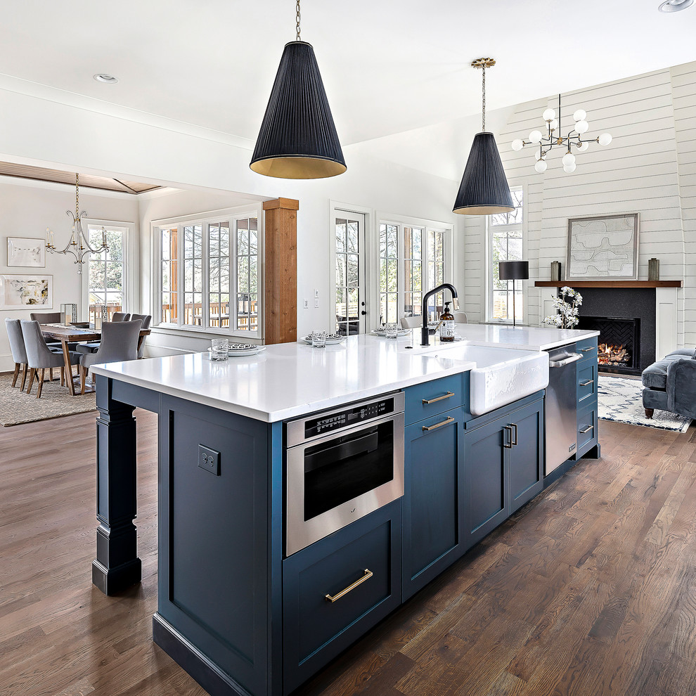 This is an example of a large country l-shaped eat-in kitchen in Charlotte with a farmhouse sink, recessed-panel cabinets, blue cabinets, quartz benchtops, white splashback, subway tile splashback, stainless steel appliances, medium hardwood floors, with island, brown floor and white benchtop.