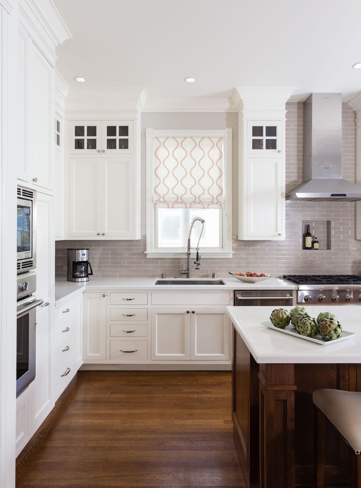 This is an example of a large traditional l-shaped eat-in kitchen in San Francisco with an undermount sink, shaker cabinets, white cabinets, solid surface benchtops, grey splashback, subway tile splashback, stainless steel appliances, dark hardwood floors, with island, brown floor and white benchtop.