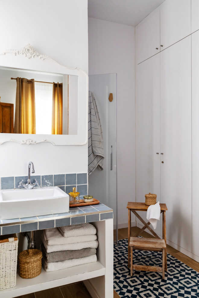 This is an example of an eclectic bathroom in Madrid with open cabinets, white cabinets, an alcove shower, white walls, a vessel sink, tile benchtops, multi-coloured floor, a hinged shower door, grey benchtops and a single vanity.