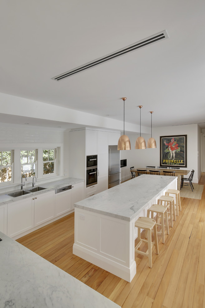 Large traditional l-shaped eat-in kitchen in Sydney with a double-bowl sink, shaker cabinets, white cabinets, marble benchtops, white splashback, ceramic splashback, stainless steel appliances, light hardwood floors, with island and yellow floor.