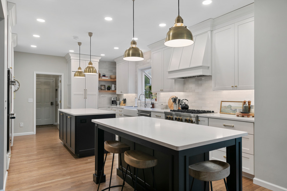 Design ideas for a transitional u-shaped kitchen in St Louis with a farmhouse sink, shaker cabinets, white cabinets, white splashback, stainless steel appliances, medium hardwood floors, multiple islands, brown floor and white benchtop.