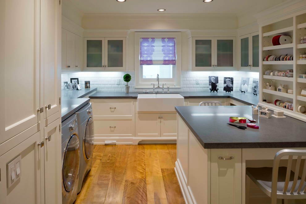 This is an example of a mid-sized transitional u-shaped dedicated laundry room in Los Angeles with a farmhouse sink, glass-front cabinets, white cabinets, soapstone benchtops, white walls, medium hardwood floors and a side-by-side washer and dryer.