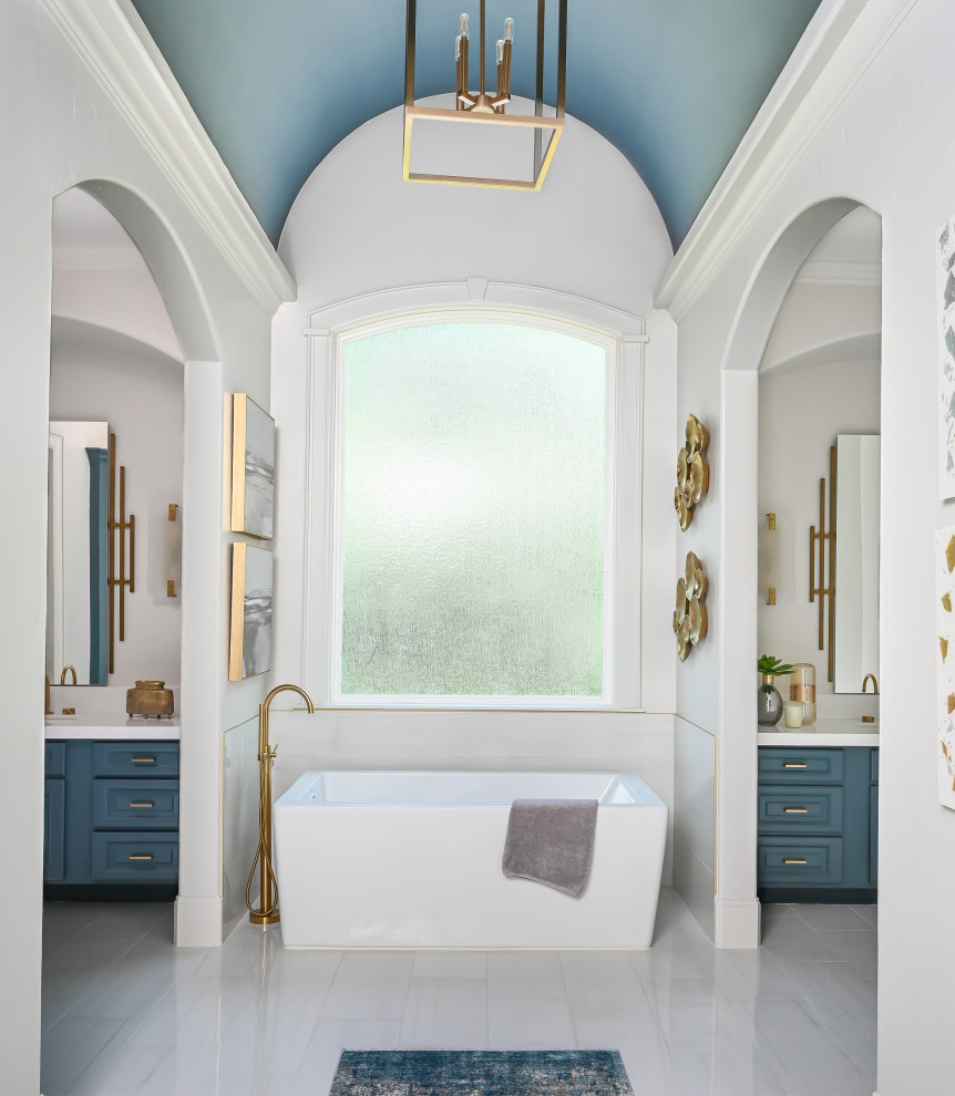 На фото: большая главная ванная комната в стиле неоклассика (современная классика) с фасадами с выступающей филенкой, синими фасадами, отдельно стоящей ванной, белыми стенами, полом из керамогранита, накладной раковиной, столешницей из кварцита, белым полом, белой столешницей, тумбой под одну раковину, встроенной тумбой и сводчатым потолком
