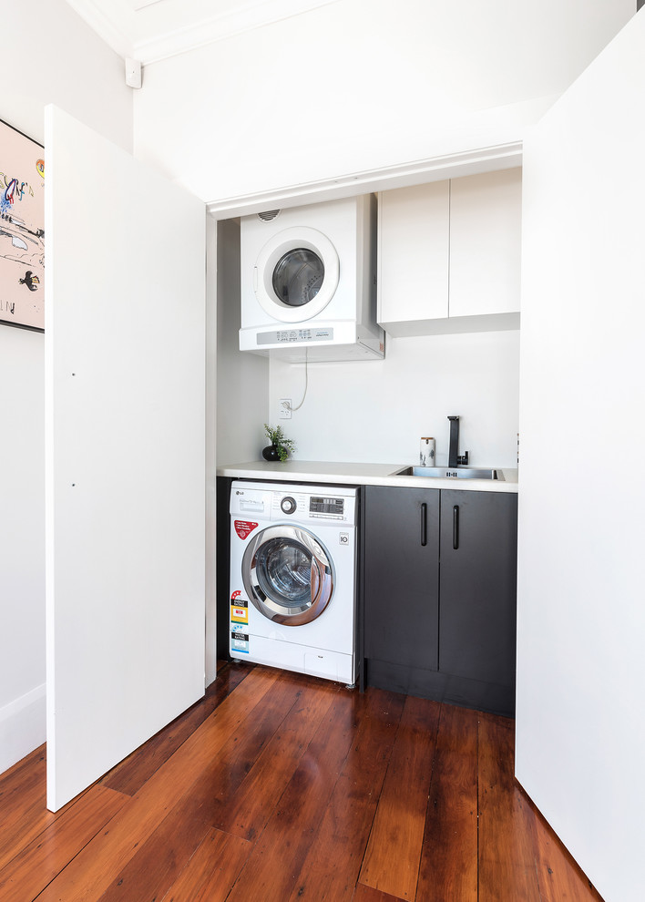 This is an example of a mid-sized modern u-shaped laundry room in Auckland with an undermount sink, flat-panel cabinets, black cabinets, laminate benchtops, white splashback, ceramic splashback, medium hardwood floors and brown floor.