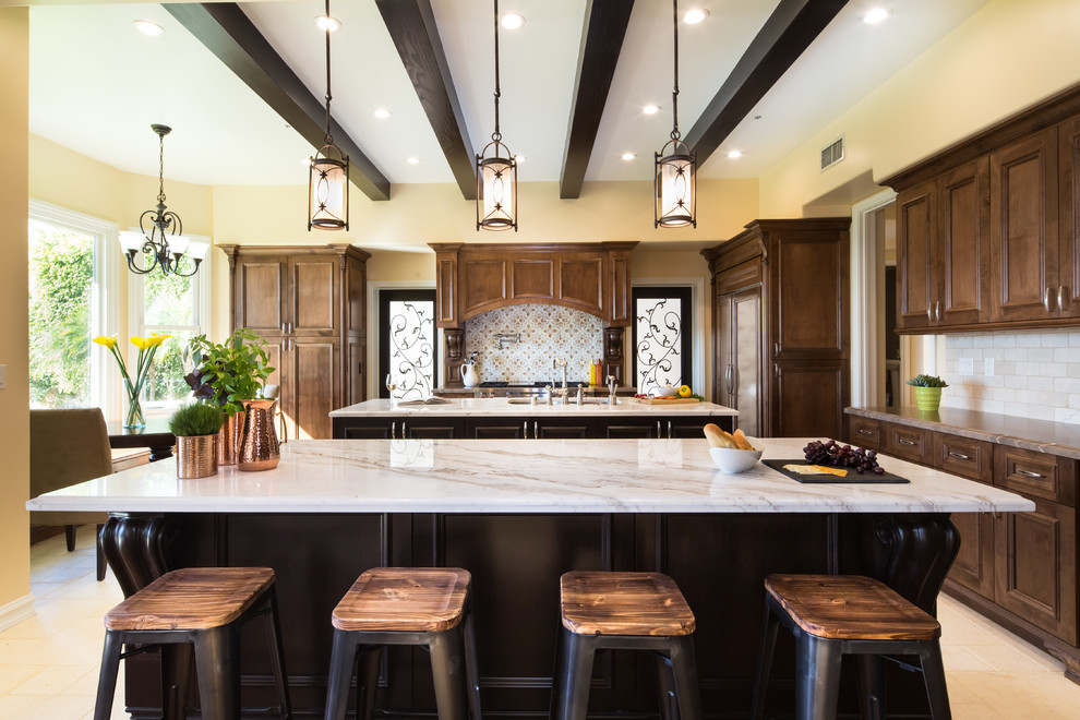 Expansive traditional eat-in kitchen in Los Angeles with an undermount sink, recessed-panel cabinets, dark wood cabinets, marble benchtops, beige splashback, stone tile splashback, stainless steel appliances, travertine floors, multiple islands and beige floor.