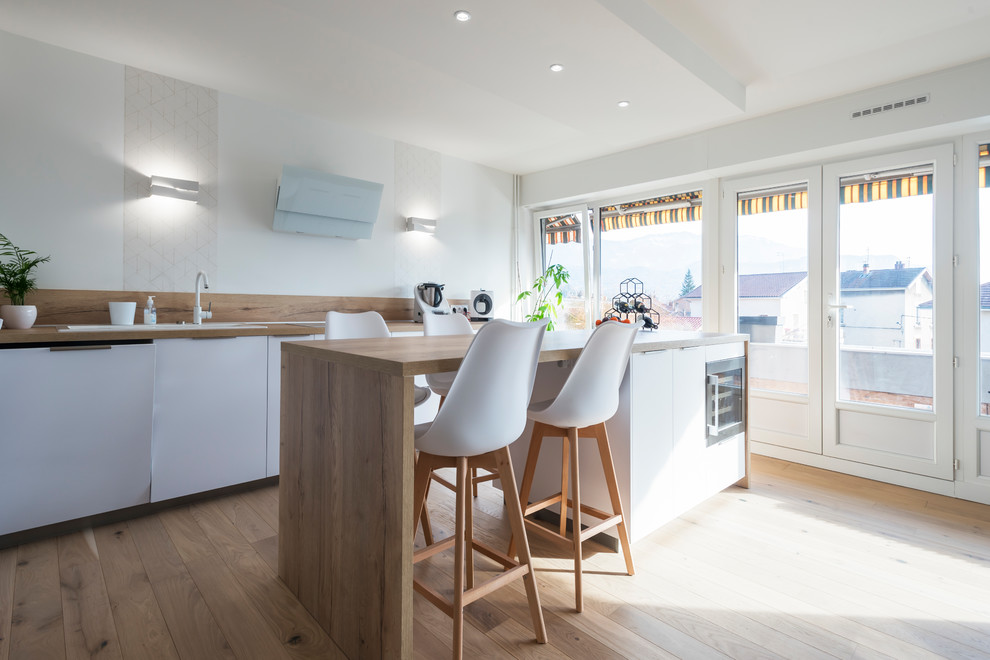 Large scandinavian galley open plan kitchen in Grenoble with a double-bowl sink, recessed-panel cabinets, white cabinets, wood benchtops, beige splashback, timber splashback, panelled appliances, light hardwood floors, with island, beige floor and beige benchtop.