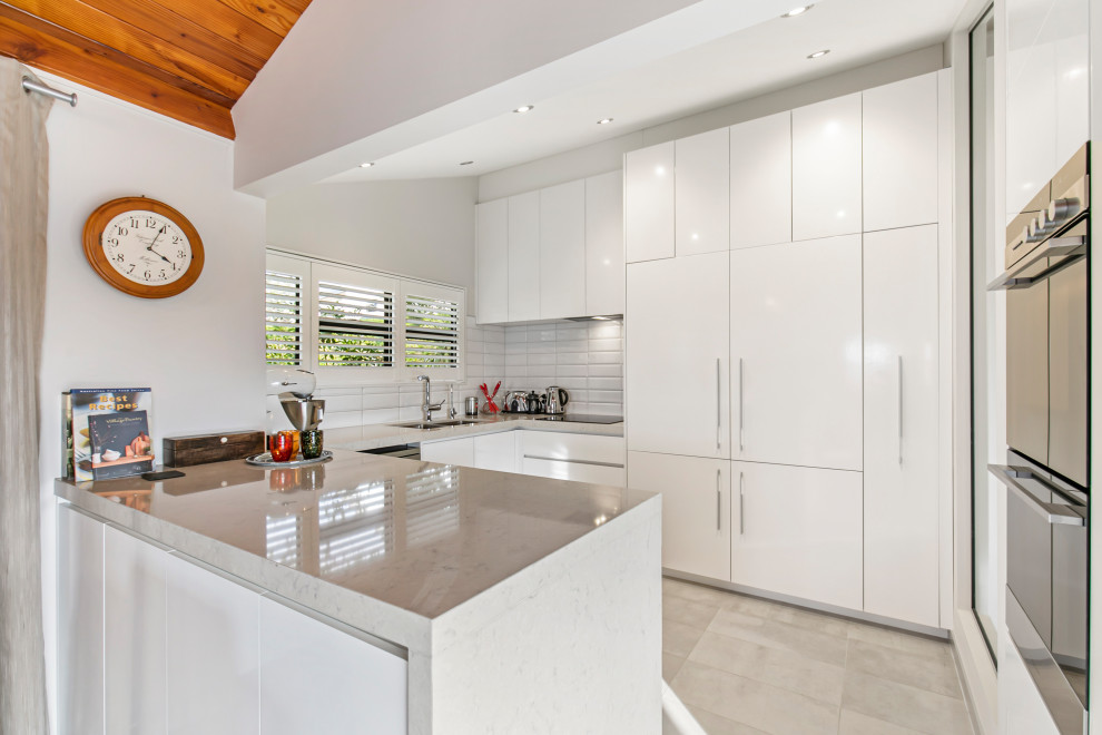 This is an example of a contemporary u-shaped kitchen in Auckland with a double-bowl sink, white cabinets, white splashback, subway tile splashback, porcelain floors, a peninsula, white floor and white benchtop.