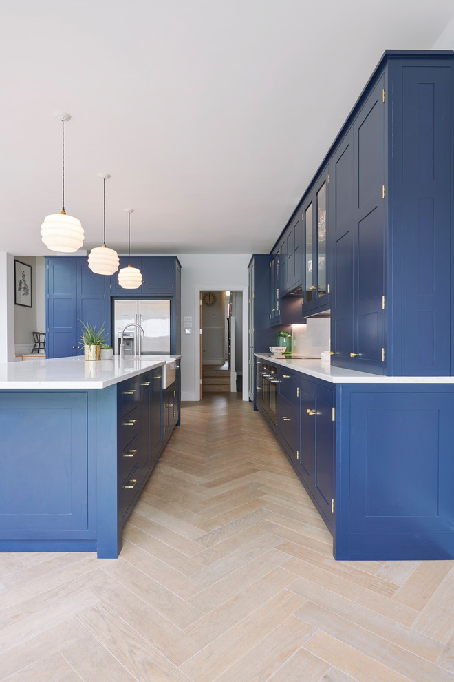 This is an example of a mid-sized modern single-wall open plan kitchen in London with a farmhouse sink, shaker cabinets, blue cabinets, quartzite benchtops, white splashback, stainless steel appliances, porcelain floors, with island and white benchtop.