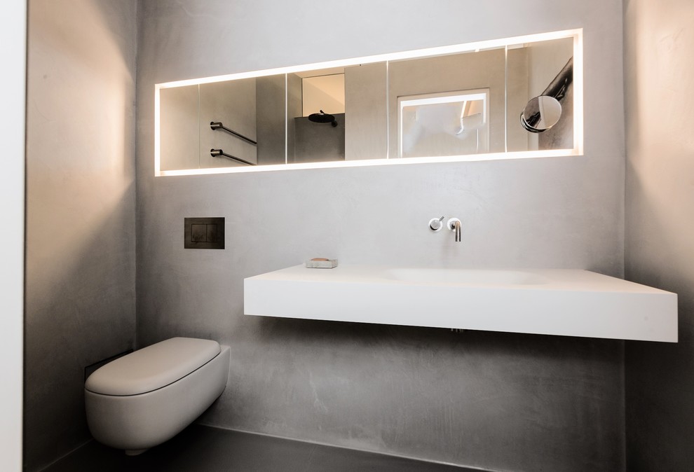 Design ideas for a small modern powder room in Munich with flat-panel cabinets, a wall-mount toilet, gray tile, grey walls, concrete floors, a trough sink, solid surface benchtops and grey floor.