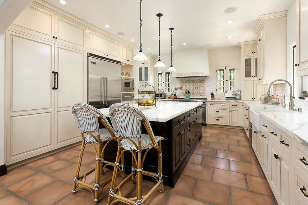 This is an example of a large mediterranean u-shaped eat-in kitchen in Los Angeles with a farmhouse sink, recessed-panel cabinets, beige cabinets, quartzite benchtops, white splashback, subway tile splashback, stainless steel appliances, terra-cotta floors, with island and brown floor.