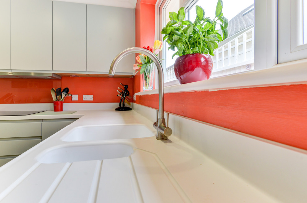 サセックスにあるお手頃価格の中くらいなビーチスタイルのおしゃれなキッチン (ドロップインシンク、フラットパネル扉のキャビネット、緑のキャビネット、人工大理石カウンター、オレンジのキッチンパネル、ガラス板のキッチンパネル、黒い調理設備、リノリウムの床、白い床、白いキッチンカウンター) の写真