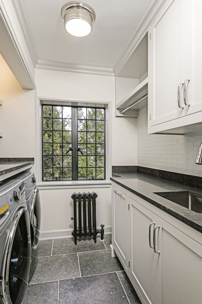 This is an example of a small traditional single-wall dedicated laundry room in New York with an undermount sink, shaker cabinets, grey cabinets, limestone benchtops, grey walls, limestone floors, a side-by-side washer and dryer, black floor and black benchtop.