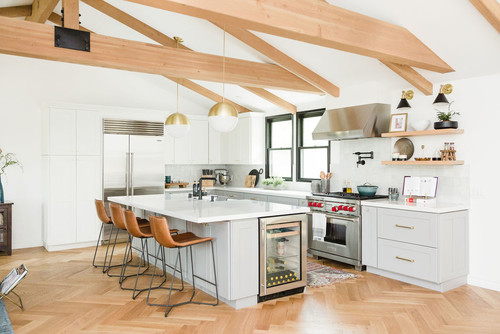 Bright kitchen with light gray Shaker base cabinets and white slab tall units, a waterfall quartz island with built-in wine fridge, brass globe pendants, exposed timber beams, and herringbone wood flooring