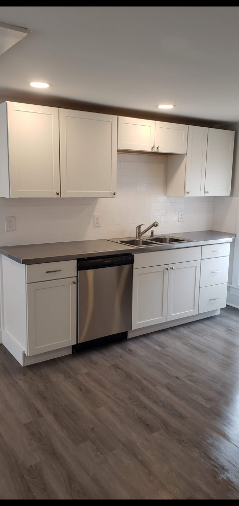 Photo of a small modern l-shaped open plan kitchen with a drop-in sink, shaker cabinets, white cabinets, stainless steel benchtops, white splashback, subway tile splashback, stainless steel appliances, vinyl floors, no island, brown floor and grey benchtop.