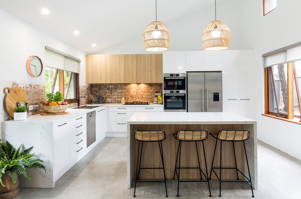 This is an example of a contemporary l-shaped kitchen in Other with a drop-in sink, flat-panel cabinets, white cabinets, brown splashback, brick splashback, stainless steel appliances, concrete floors, a peninsula, grey floor and white benchtop.