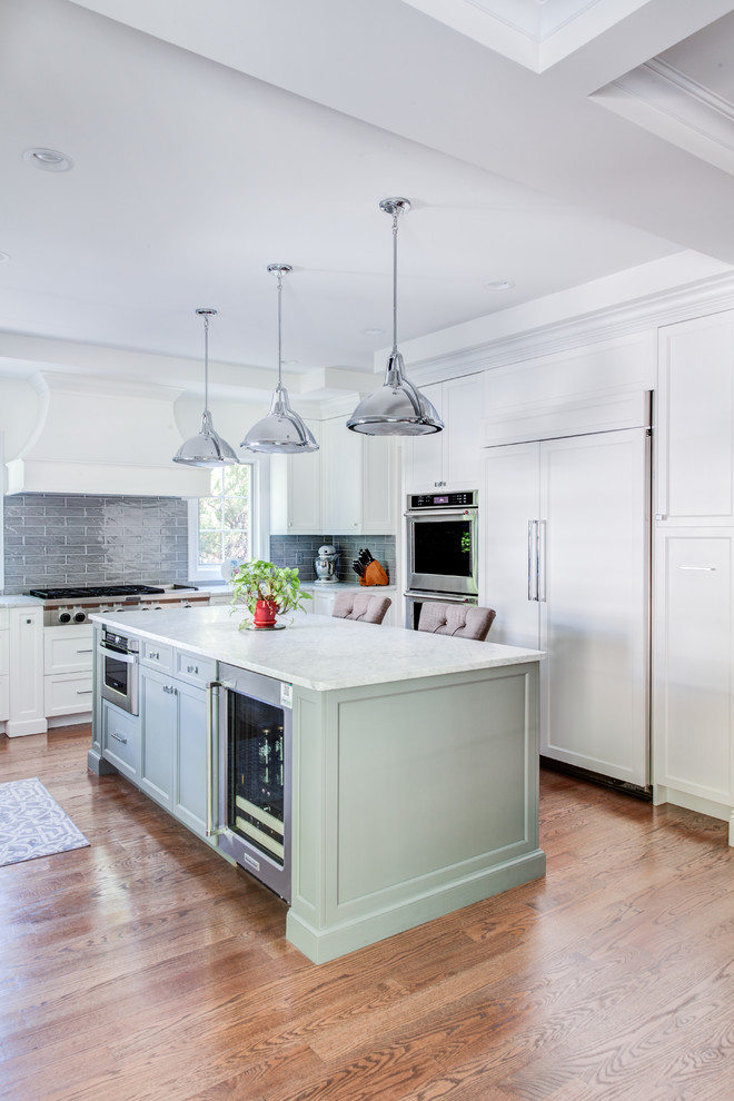 This is an example of a mid-sized transitional u-shaped open plan kitchen in New York with a farmhouse sink, shaker cabinets, white cabinets, quartz benchtops, grey splashback, glass tile splashback, panelled appliances, light hardwood floors, with island and brown floor.