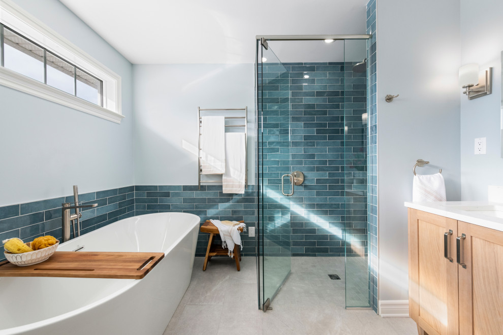 Photo of a large beach style master bathroom in Ottawa with light wood cabinets, a freestanding tub, a curbless shower, a two-piece toilet, blue tile, blue walls, an undermount sink, a hinged shower door, white benchtops, a double vanity, a freestanding vanity, shaker cabinets, ceramic tile, ceramic floors, engineered quartz benchtops, blue floor and a niche.
