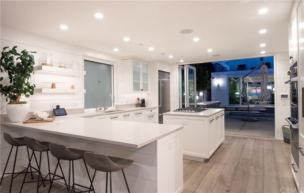 Photo of a small contemporary l-shaped eat-in kitchen in Los Angeles with a single-bowl sink, shaker cabinets, white cabinets, quartz benchtops, engineered quartz splashback, stainless steel appliances, medium hardwood floors, with island and grey floor.