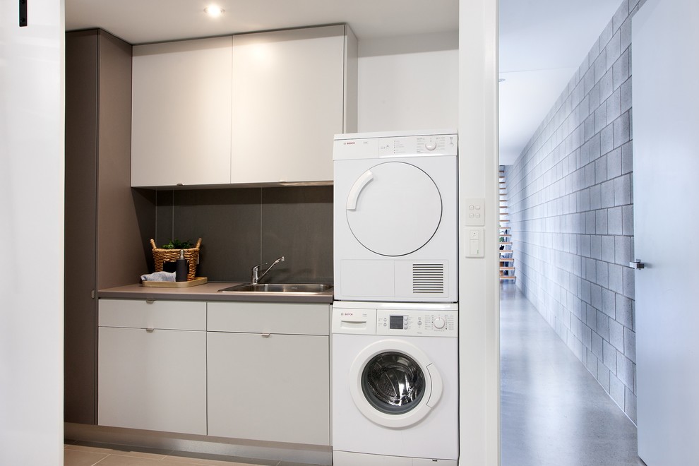 Photo of a mid-sized modern single-wall dedicated laundry room in Sunshine Coast with flat-panel cabinets, a single-bowl sink, white cabinets, laminate benchtops, white walls, ceramic floors and a stacked washer and dryer.