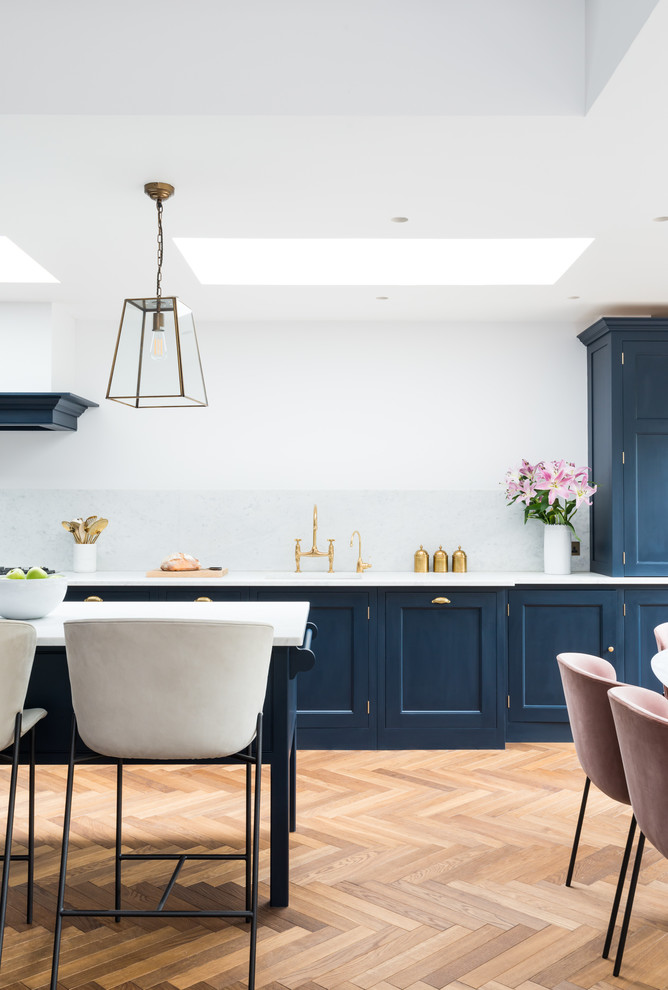 Photo of an expansive transitional single-wall eat-in kitchen in London with a drop-in sink, recessed-panel cabinets, blue cabinets, marble benchtops, white splashback, marble splashback, white appliances, light hardwood floors, a peninsula and white benchtop.