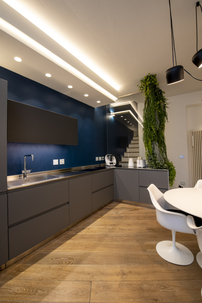 Mid-sized modern l-shaped open plan kitchen in Bologna with flat-panel cabinets, grey cabinets, stainless steel benchtops, blue splashback and no island.