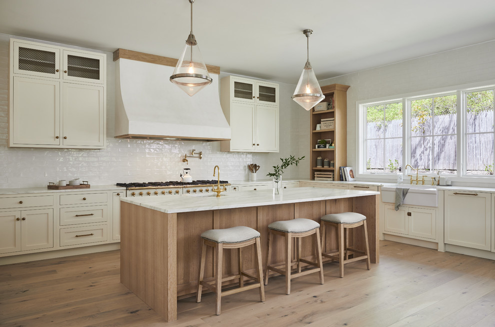 Inspiration for a country l-shaped kitchen in New York with a farmhouse sink, shaker cabinets, white cabinets, white splashback, white appliances, medium hardwood floors, with island, brown floor and white benchtop.