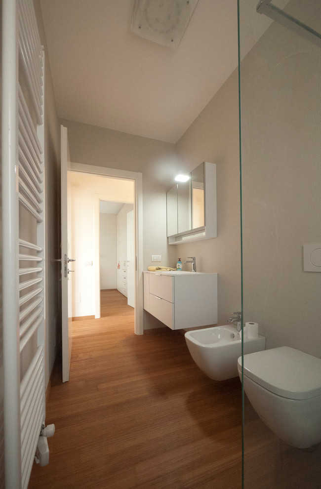 This is an example of a mid-sized modern master bathroom in Milan with an integrated sink, flat-panel cabinets, white cabinets, engineered quartz benchtops, an open shower, a wall-mount toilet, blue tile, glass tile, beige walls and bamboo floors.