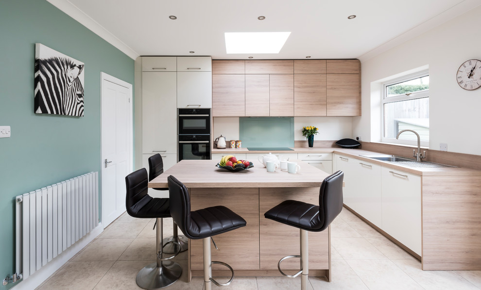 This is an example of a mid-sized contemporary l-shaped eat-in kitchen in Surrey with a single-bowl sink, flat-panel cabinets, light wood cabinets, laminate benchtops, green splashback, glass sheet splashback, black appliances, ceramic floors, with island, beige floor and beige benchtop.