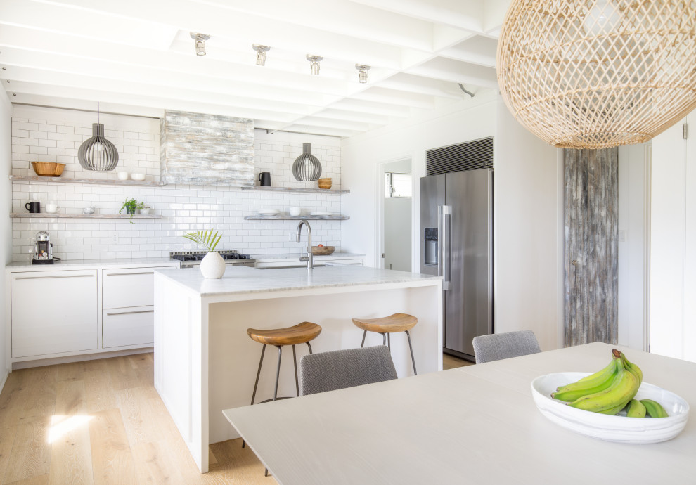 This is an example of a beach style galley kitchen in Other with a farmhouse sink, flat-panel cabinets, white cabinets, white splashback, subway tile splashback, stainless steel appliances, light hardwood floors, with island, beige floor, white benchtop and exposed beam.