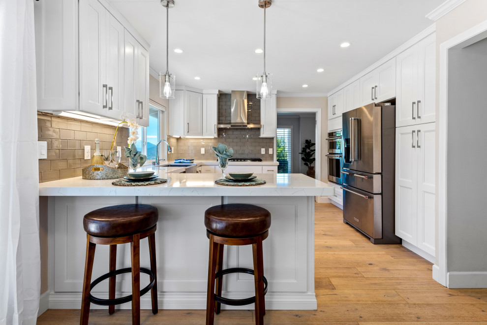Inspiration for a large transitional u-shaped medium tone wood floor and brown floor eat-in kitchen remodel with a farmhouse sink, recessed-panel cabinets, white cabinets, gray backsplash, stainless steel appliances, a peninsula and white countertops