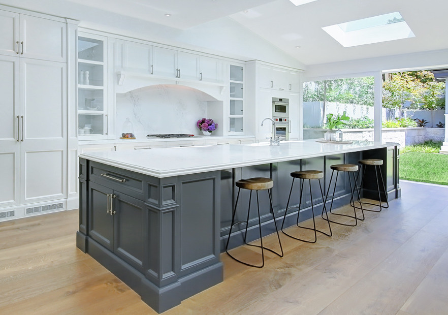 This is an example of a large contemporary galley kitchen pantry in Sydney with a belfast sink, shaker cabinets, white cabinets, engineered stone countertops, white splashback, engineered quartz splashback, stainless steel appliances, light hardwood flooring, an island, brown floors and white worktops.