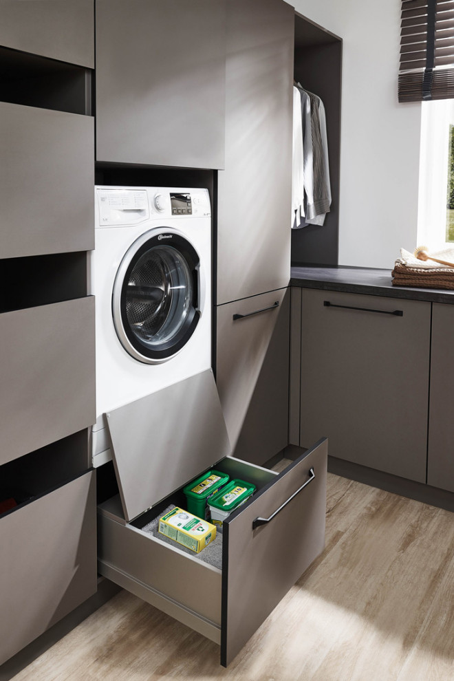 Modelo de armario lavadero en L escandinavo de tamaño medio con armarios con paneles lisos, puertas de armario grises, encimera de laminado, paredes blancas, suelo laminado, lavadora y secadora integrada, suelo beige y encimeras grises