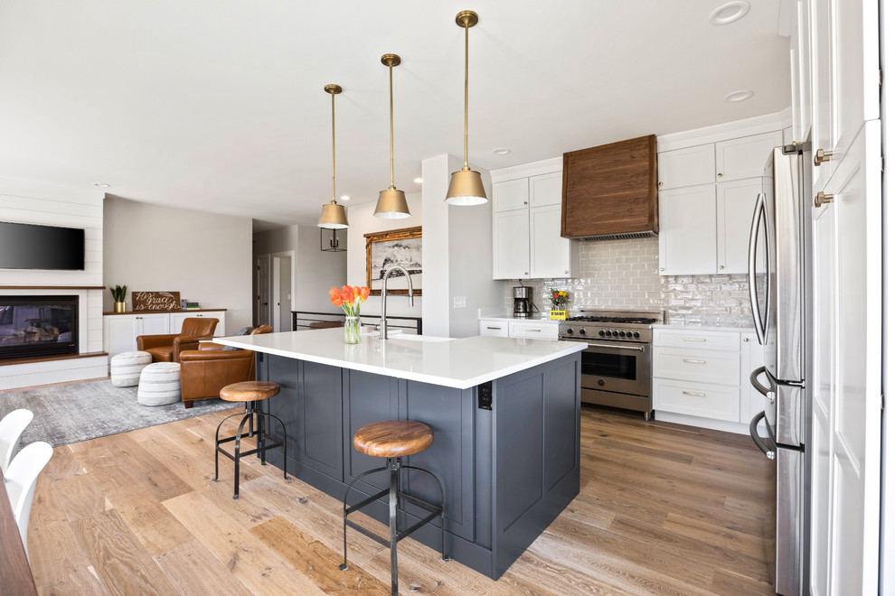 Example of a transitional l-shaped light wood floor and brown floor kitchen design with a farmhouse sink, shaker cabinets, white cabinets, quartz countertops, white backsplash, subway tile backsplash, stainless steel appliances, an island and white countertops