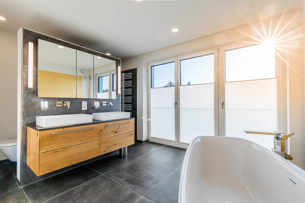 Design ideas for a contemporary master bathroom in Nuremberg with flat-panel cabinets, medium wood cabinets, a freestanding tub, white walls, a vessel sink, grey floor and black benchtops.