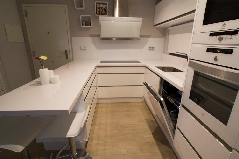 This is an example of a small contemporary u-shaped open plan kitchen in Malaga with a single-bowl sink, flat-panel cabinets, white cabinets, white splashback, white appliances, light hardwood floors and no island.