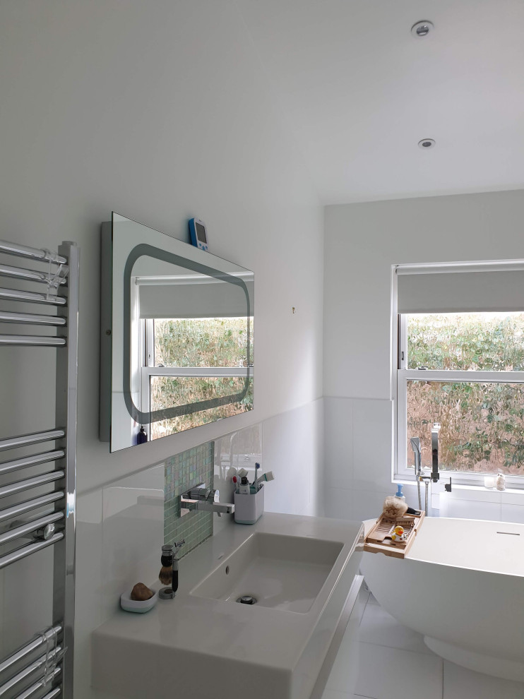 This is an example of a large modern kids bathroom in London with flat-panel cabinets, white cabinets, a freestanding tub, an open shower, a wall-mount toilet, white tile, porcelain tile, white walls, pebble tile floors, a drop-in sink, marble benchtops, white floor, an open shower, white benchtops, a single vanity, a built-in vanity and recessed.